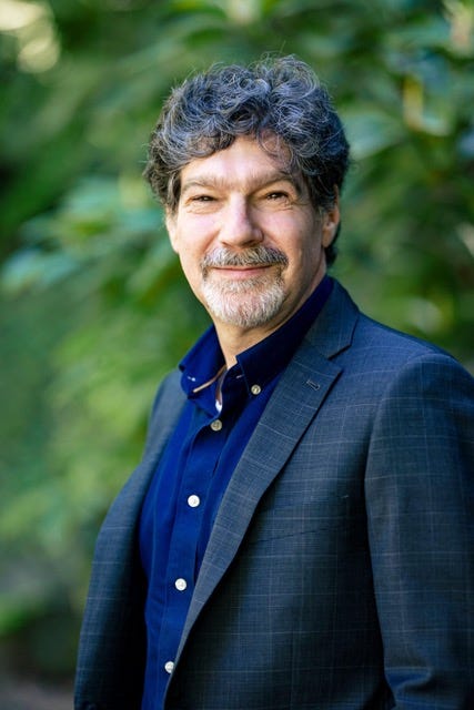 Photograph of a smiling middle-aged white man wearing a blue button-down shirt and blazer in front of a green forest background.