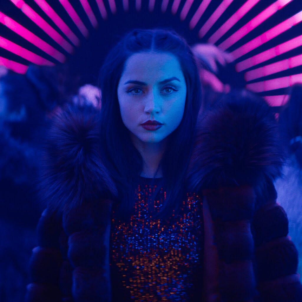 A woman wearing a fur coat stands in the center of a neon-lit space with pink and blue lights, surrounded by people dancing in the background. She looks directly at the camera with a confident expression.