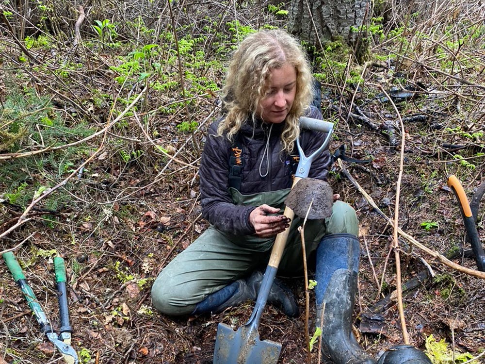 A woman with long, curly, blond hair wears rubber overalls and holds a spade in a forested setting. A woman with long, curly, blond hair wears rubber overalls and holds a spade in a forested setting.