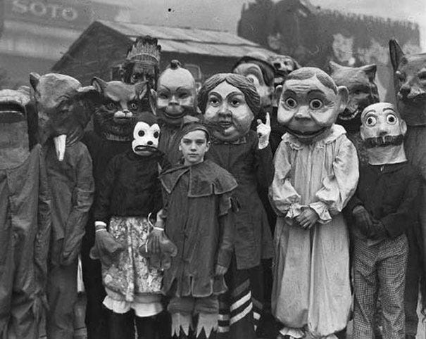 Old black and white photo of a group of people in masks--a homemade mickey mouse, cats, foxes and giant babies among others. A boy at the centre is unmaked in a medeival tunic, staring directly at the camera. 