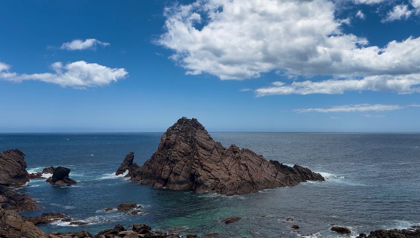 Wide coastal view of Sugarloaf Rock rising from the Indian Ocean, with calm blue water, a pale horizon, and scattered clouds in a bright sky.