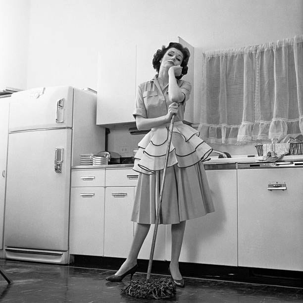 1950s DAYDREAMING BORED WOMAN HOUSEWIFE WEARING APRON LEANING ON MOP WHILE CLEANING KITCHEN FLOOR