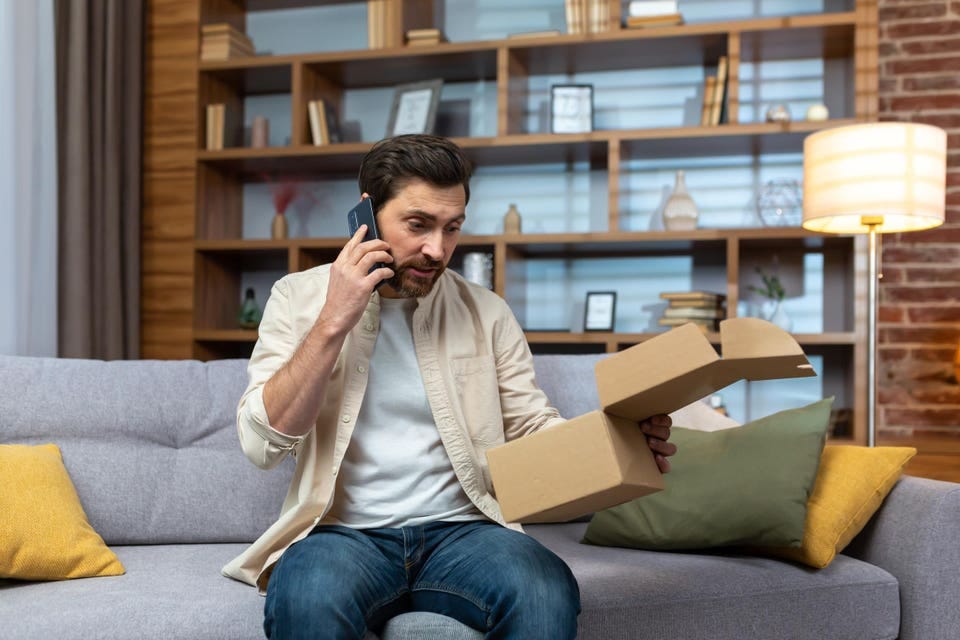An angry young man is sitting at home on the couch, holding an open box in his hands. I received a wrong, empty, damaged parcel. Talks on the phone with the delivery service, the store