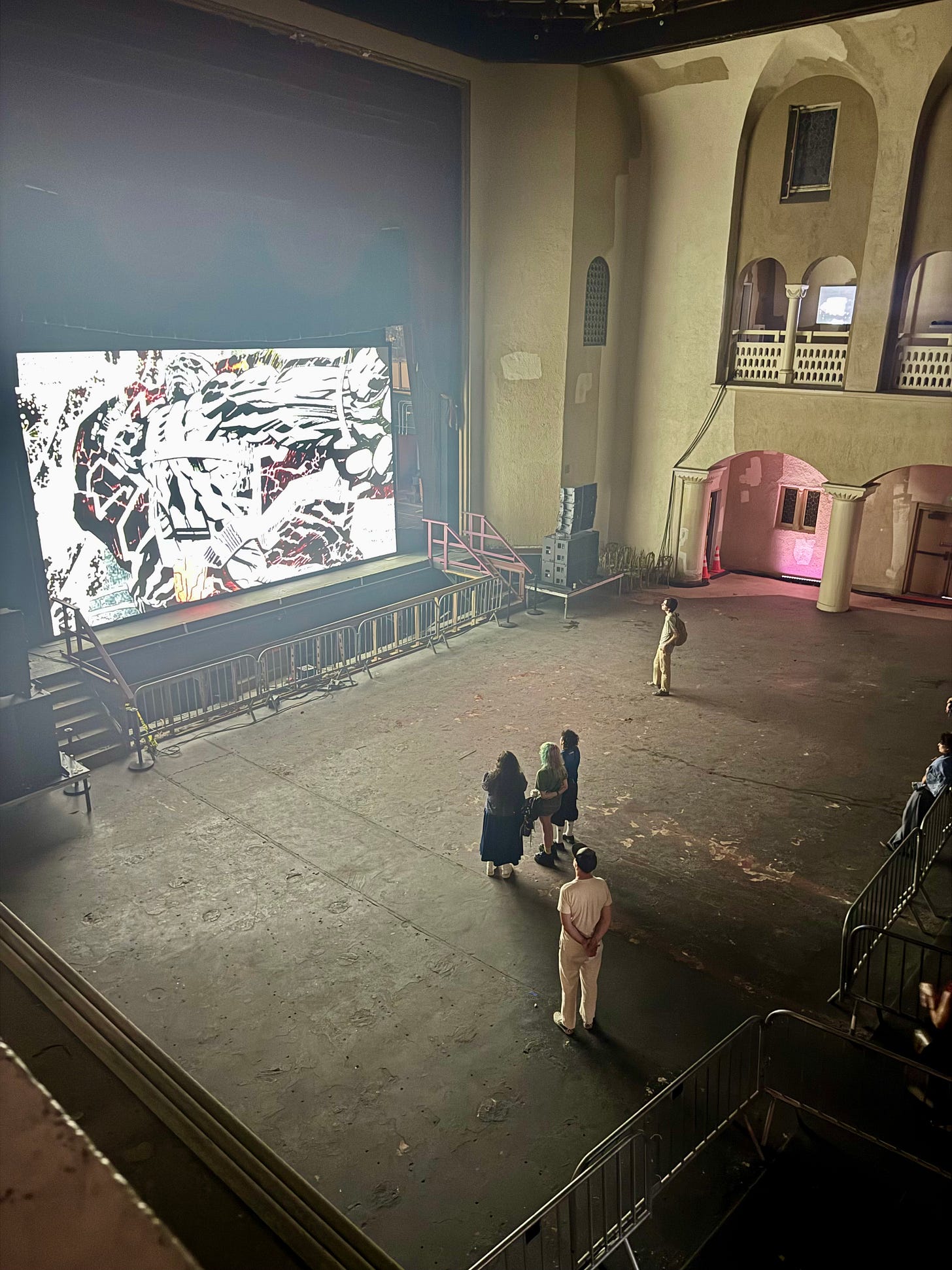 View from the balcony of the Variety Arts Theater main auditorium showing visitors standing on the worn      concrete floor looking up at a large black and white video projection on the stage with ornate arched walls and          balustrades visible  