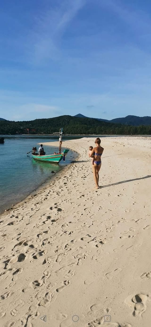 A young woman in a bikini carrying a small child on her hip walks down a sandy beach towards a green fishing boat against a blue sky with mountains in the distance.