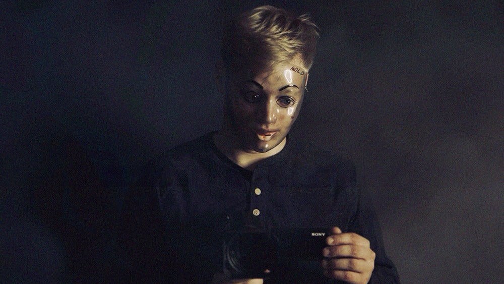 The character Milk from 'Milk & Serial' wearing a transparent plastic full-face mask and looking at the screen of a video camera