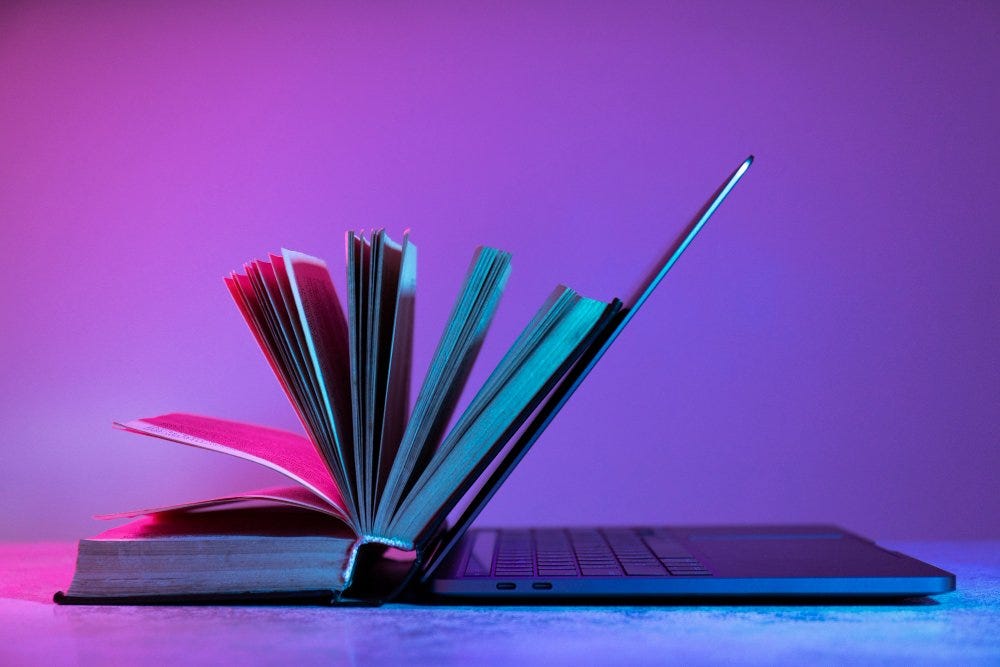 #descrevemospravocê  A imagem mostra um livro aberto encostado na tela de um notebook semiaberto, formando juntos um ângulo simétrico. O livro está com as páginas levemente viradas para cima, como se estivessem sendo folheadas pelo vento, e sua capa é dura, de cor escura. O notebook, posicionado à direita, tem design moderno e fino, com a tela levantada, apoiando o livro. O fundo é um degradê vibrante em tons de roxo, rosa e azul, com iluminação artificial que também colore o livro e o computador nessas mesmas tonalidades. A imagem transmite a ideia de conexão entre o conhecimento tradicional, representado pelo livro, e o digital, simbolizado pelo notebook.