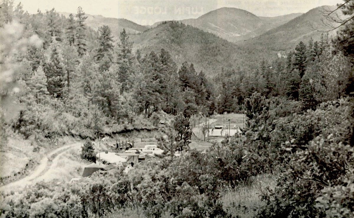 Black-and-white aerial photograph of the Colorado Sunshine Club from the 1950s, showing rustic buildings, parked cars, and tennis courts surrounded by dense pine forest and mountain peaks in the background. Black-and-white aerial photograph of the Colorado Sunshine Club from the 1950s, showing rustic buildings, parked cars, and tennis courts surrounded by dense pine forest and mountain peaks in the background.