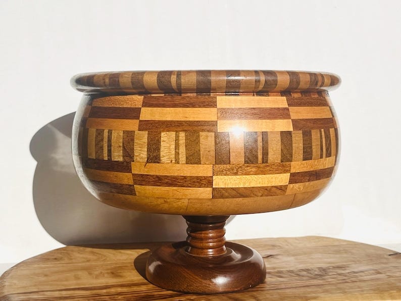 May include: A wooden bowl with a pedestal base. The bowl is made of various shades of wood, creating a checkered pattern. The bowl is brown and has a smooth, polished finish.