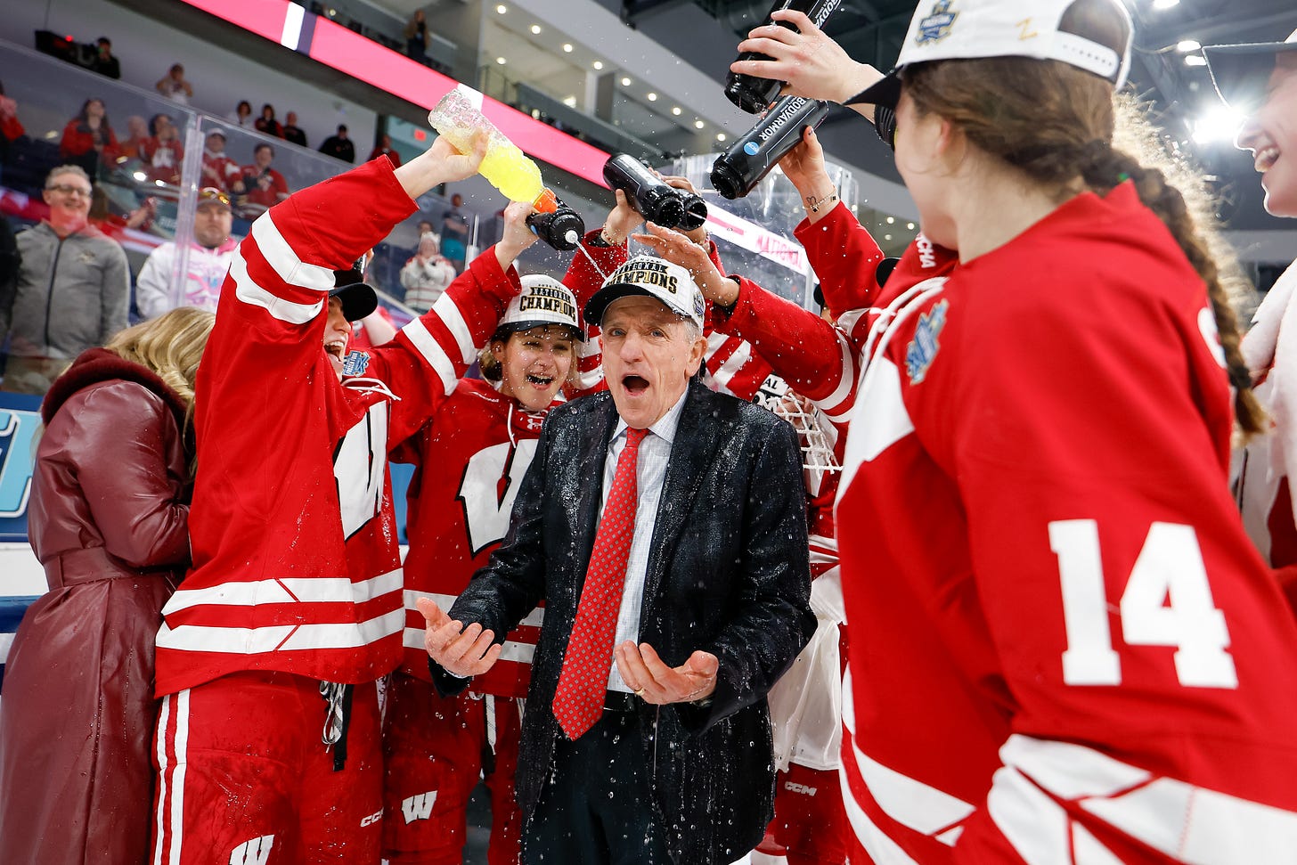 Mark Johnson, wearing a black suit with a red tie, is surrounded by Wisconsin hockey players who squirt liquid on his head using water bottles