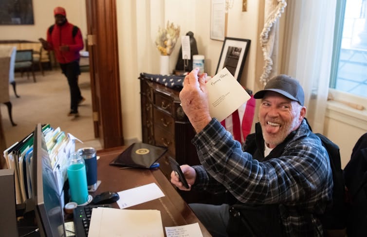 Richard Barnett sits at a desk holding a piece of mail with Nancy Pelosi's name and sticking his tongue out. Richard Barnett sits at a desk holding a piece of mail with Nancy Pelosi's name and sticking his tongue out.