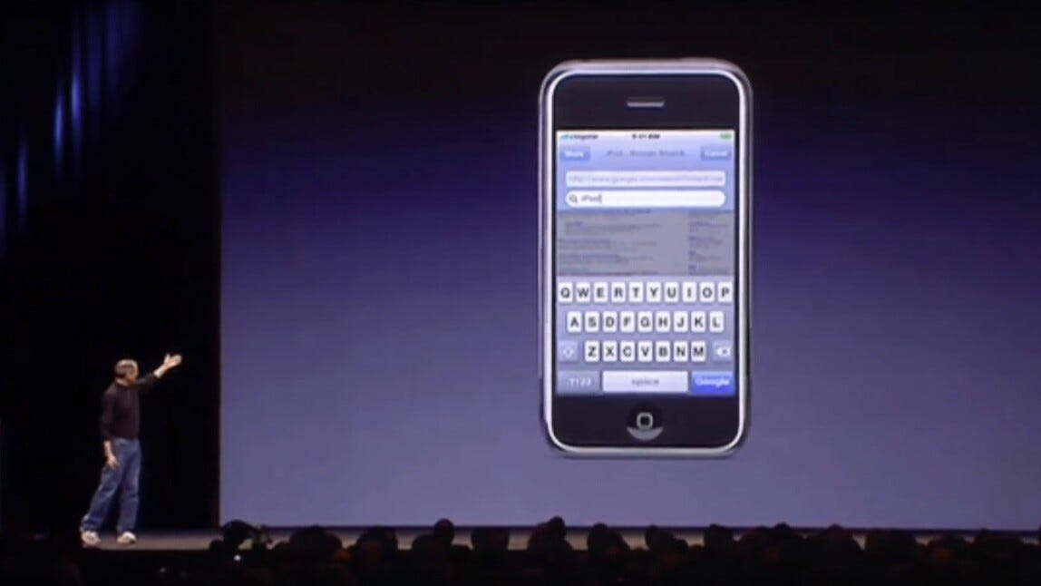 Photograph from January 9, 2007 showing Steve Jobs on stage at the Macworld keynote presentation, gesturing toward a large screen displaying the original iPhone with its on-screen keyboard visible. Jobs is wearing his signature black turtleneck and jeans, standing in front of an auditorium audience. The image captures the historic moment when Apple launched the iPhone, igniting a global race to build mobile applications. While headlines celebrated viral app successes like Instagram's billion-dollar acquisition and Angry Birds, the vast majority of the 90% of apps generated less than $5,000 in lifetime revenue. The true winners were infrastructure providers—ARM, Qualcomm, Samsung—who sold processors, modems, memory chips, and displays to every smartphone manufacturer indiscriminately, capturing far more value than those competing in the application layer.