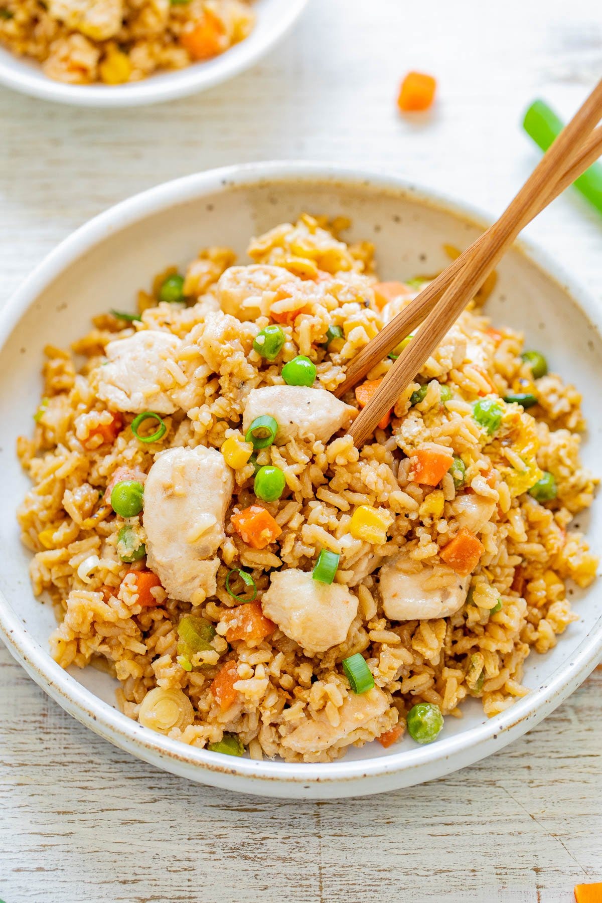 A bowl of chicken fried rice with peas, carrots, and corn, garnished with green onions. Chopsticks are placed on top of the dish.