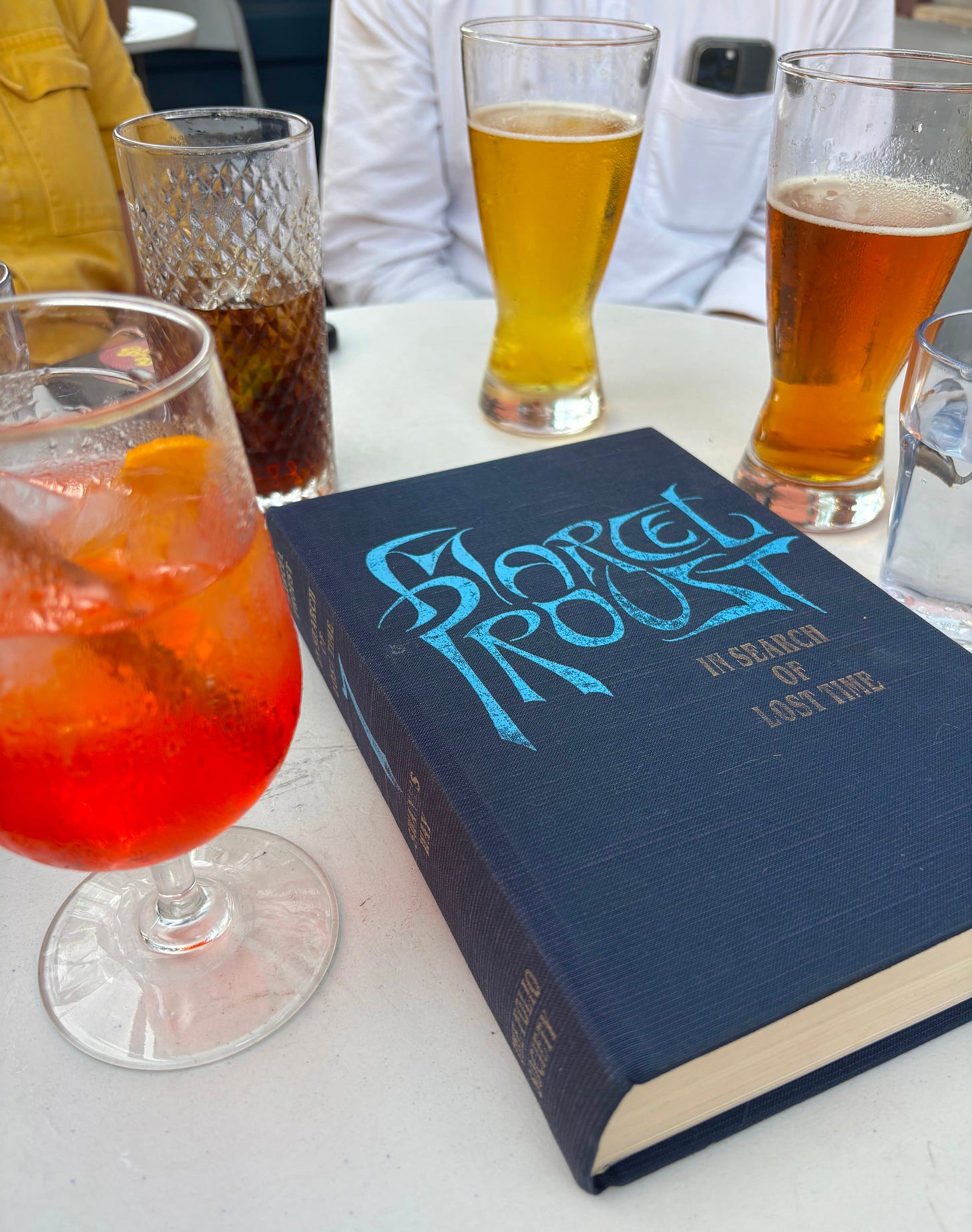 Blue hardcover of Marcel Proust's In Search of Lost Time on a white table surrounded by full drink glassses