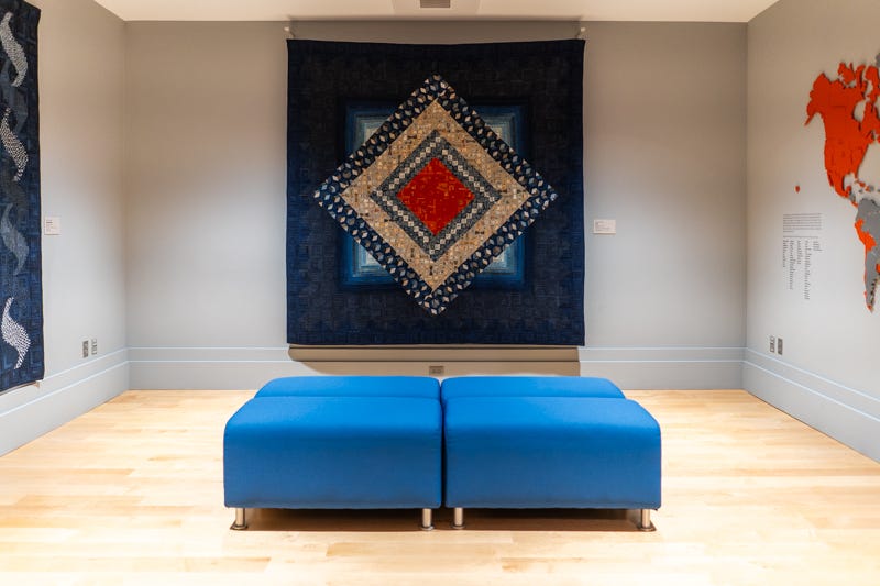 A photograph of a room in a museum showcasing a large quite with a dark blue border and interior patterns of varying shades of blue diamonds, a light brown diamond, and in interior dark red diamond. A set of four blue seats are on the wood floor. A photograph of a room in a museum showcasing a large quite with a dark blue border and interior patterns of varying shades of blue diamonds, a light brown diamond, and in interior dark red diamond. A set of four blue seats are on the wood floor.