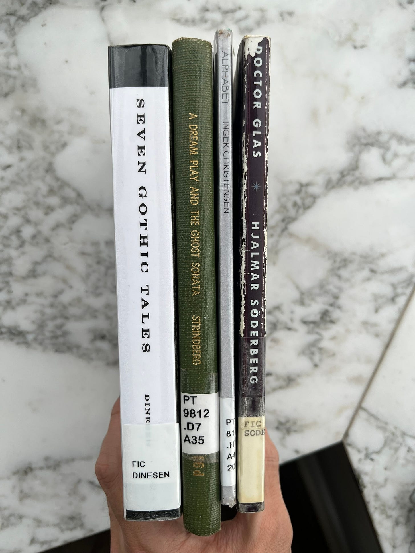 A hand holding 4 library books, with back covers showing. Back covers show, from left to right: "Seven Gothic Tales" by Dinesen, "A Dream Play and The Ghost Sonata" by Strindberg, "Alphabet" by Inger Christensen, "Doctor Glas" by Hjalmar Söderberg.