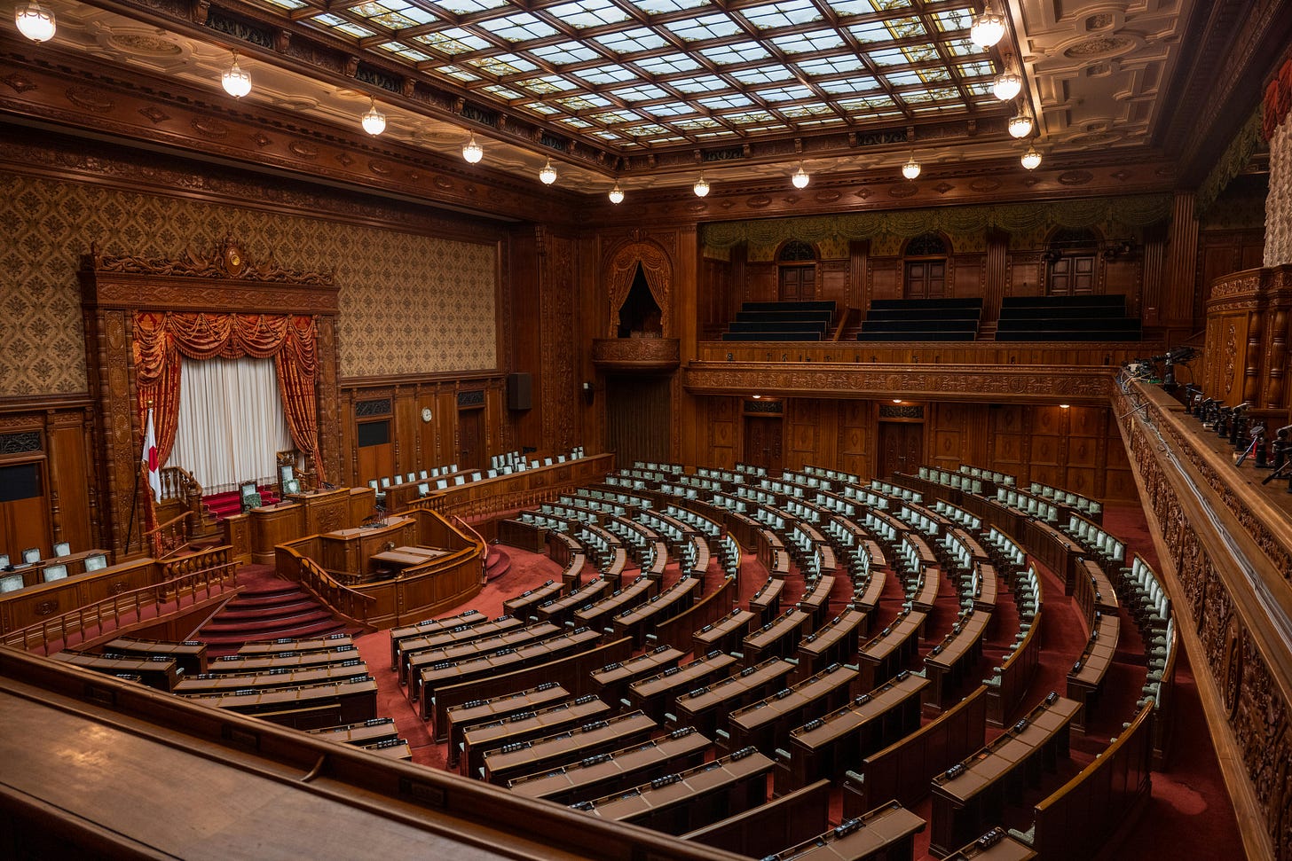 The empty wood-paneled chamber of Japan's National Diet, semicircular desks beneath a glass skylight. The empty wood-paneled chamber of Japan's National Diet, semicircular desks beneath a glass skylight.