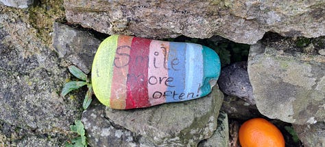 1. pebble inserted to gap between corbelled stones on the dome of the holy well that reads Jesus loves you. 2. Pebble painted in brights stripes inserted between corbelled stones that reads Smile More Often. 3. Close up of sprawling tree with large shiny green leaves with scraps of cloth, socks and all manner of oddities tied to its branches