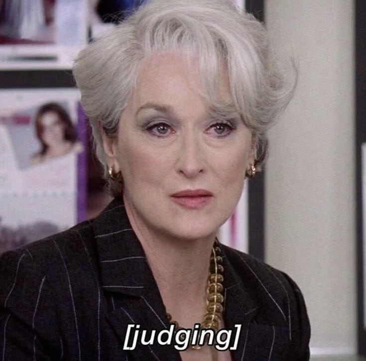 This may contain: an older woman with white hair wearing a black suit and gold necklace, looking at the camera