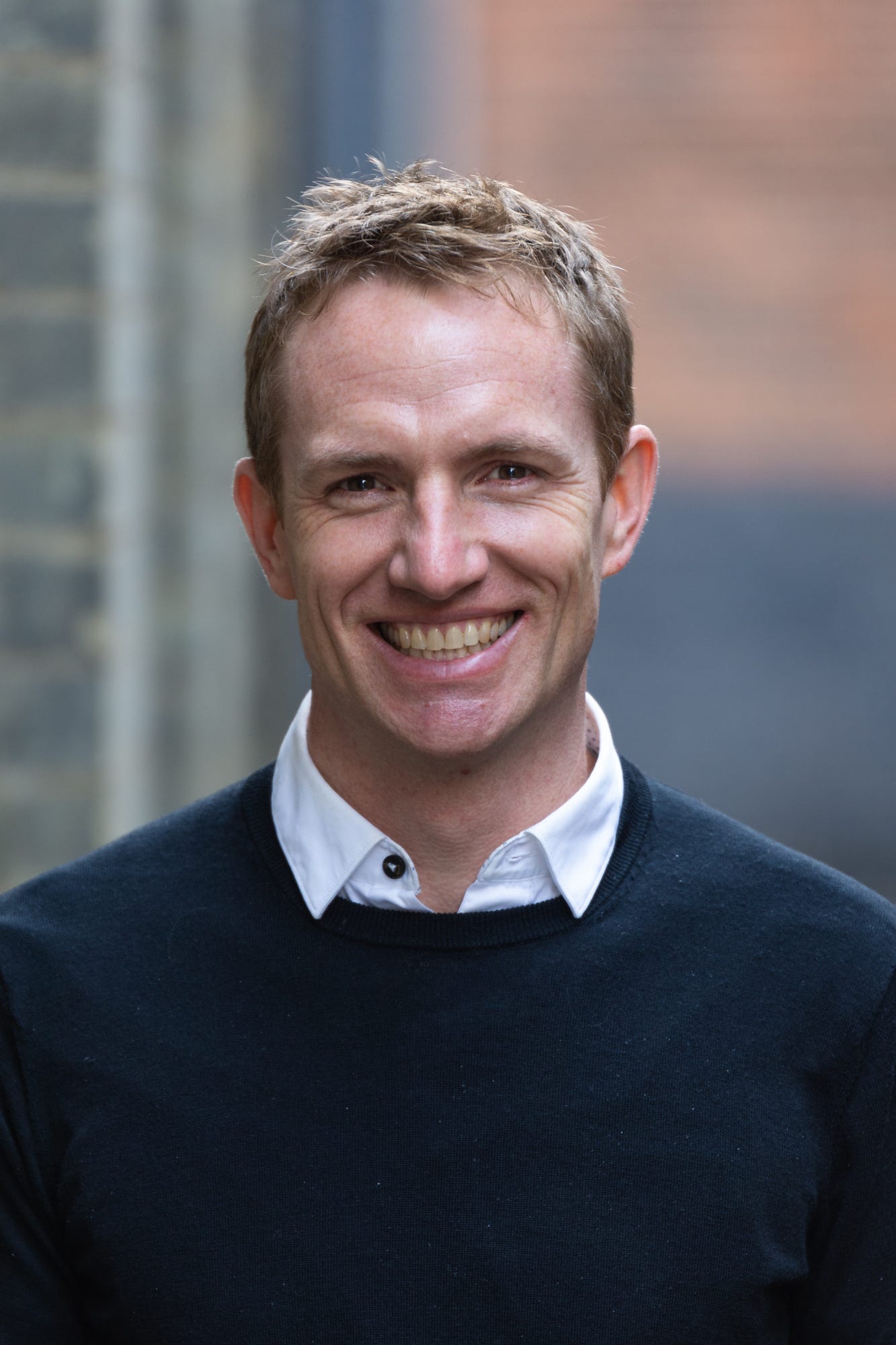 Jon is a white man with short, sandy-coloured hair. He’s wearing a plain navy crew neck sweater over white shirt with an open collar. He’s staring directly at the camera with a big open smile.