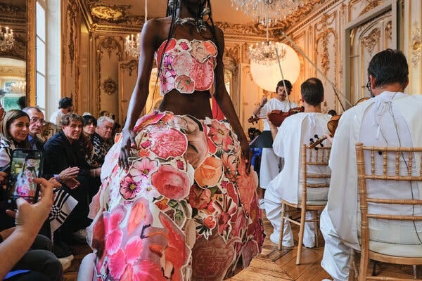 A model in an elaborate two-piece gown embellished with floral images walks close to the camera, flanked by audience members on the left and musicians in white on the right.