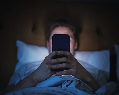 A person in bed holds a smartphone up in front of their face in dim lighting