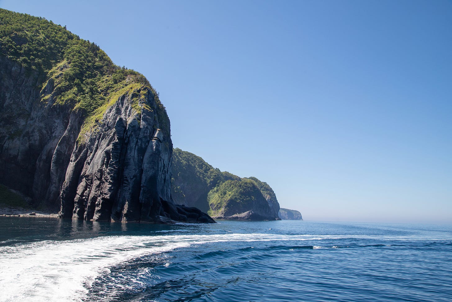 Steep green-topped sea cliffs rising from deep blue water, with a boat's wake in the foreground.