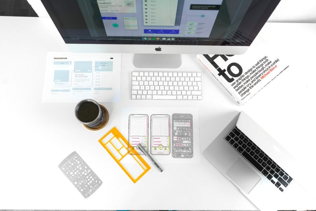 desk of a designer. a desktop computer, a laptop, coffee, a design book, and prototypes
