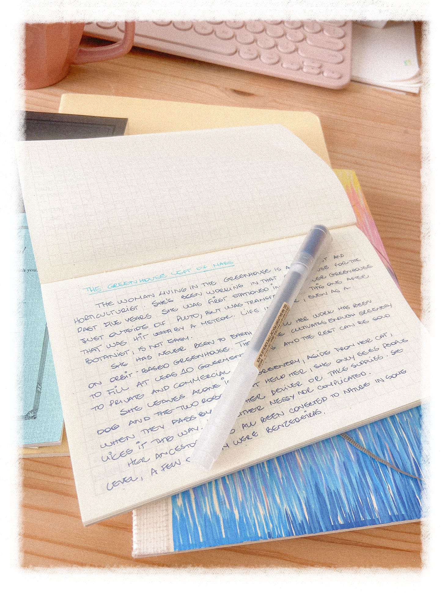 A small notebook is open over a wooden table. The paper in the notebook has a light blue grid and text is handwritten on it. Underneath the notebook, a colourful sketchbook.