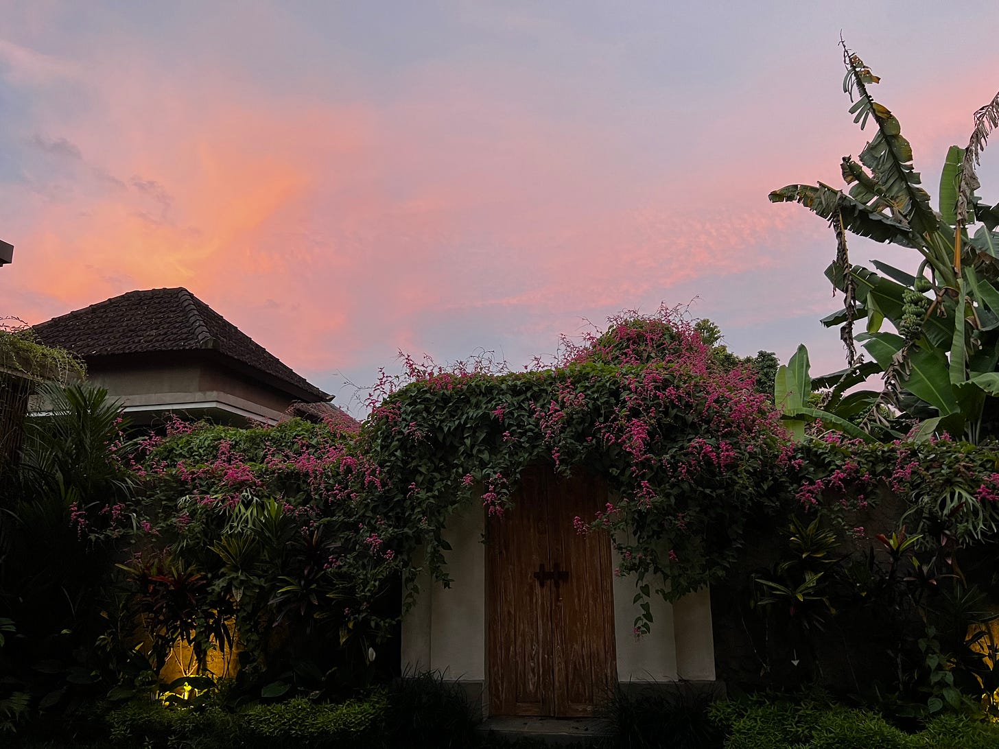 A wooden villa door at dusk, framed by a wall thick with pink bougainvillea, beneath a pink and lavender sky. A wooden villa door at dusk, framed by a wall thick with pink bougainvillea, beneath a pink and lavender sky.