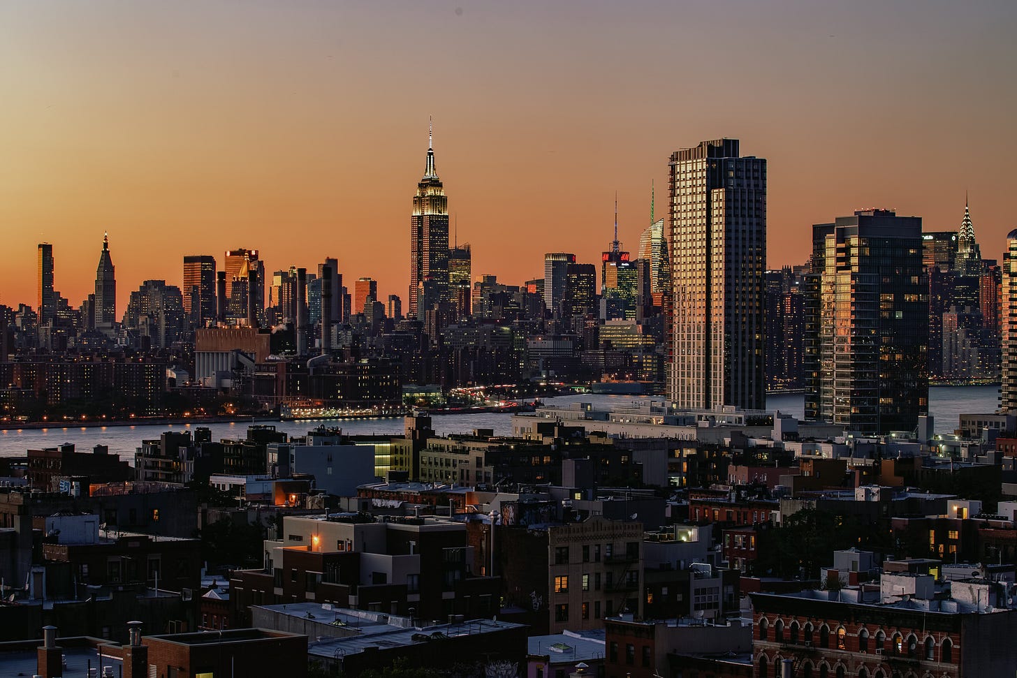 A wide view of the Midtown Manhattan skyline at sunset, seen from an elevated viewpoint in Brooklyn. The Empire State Building is prominent in the center, and the city lights are beginning to turn on against the orange sky.