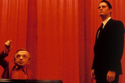 A person standing in front of a person sitting in front of a red curtain
Description automatically generated