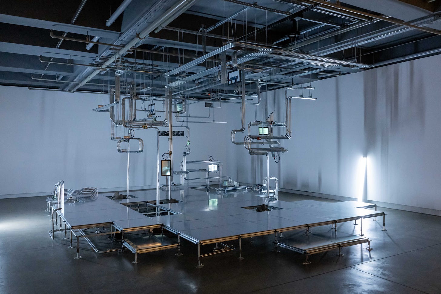 A dimly lit gallery space with spare interior fittings with a fluorescent light tube leaning against the two side walls that lights the room with an eerie blue-grey light. In the middle of the gallery, is a raised platform with a series of steel pipes, running in fixed track formation with occasional bends, that range from the ceiling to the floor in the middle of which are a number of small flat screens with graphical displays.