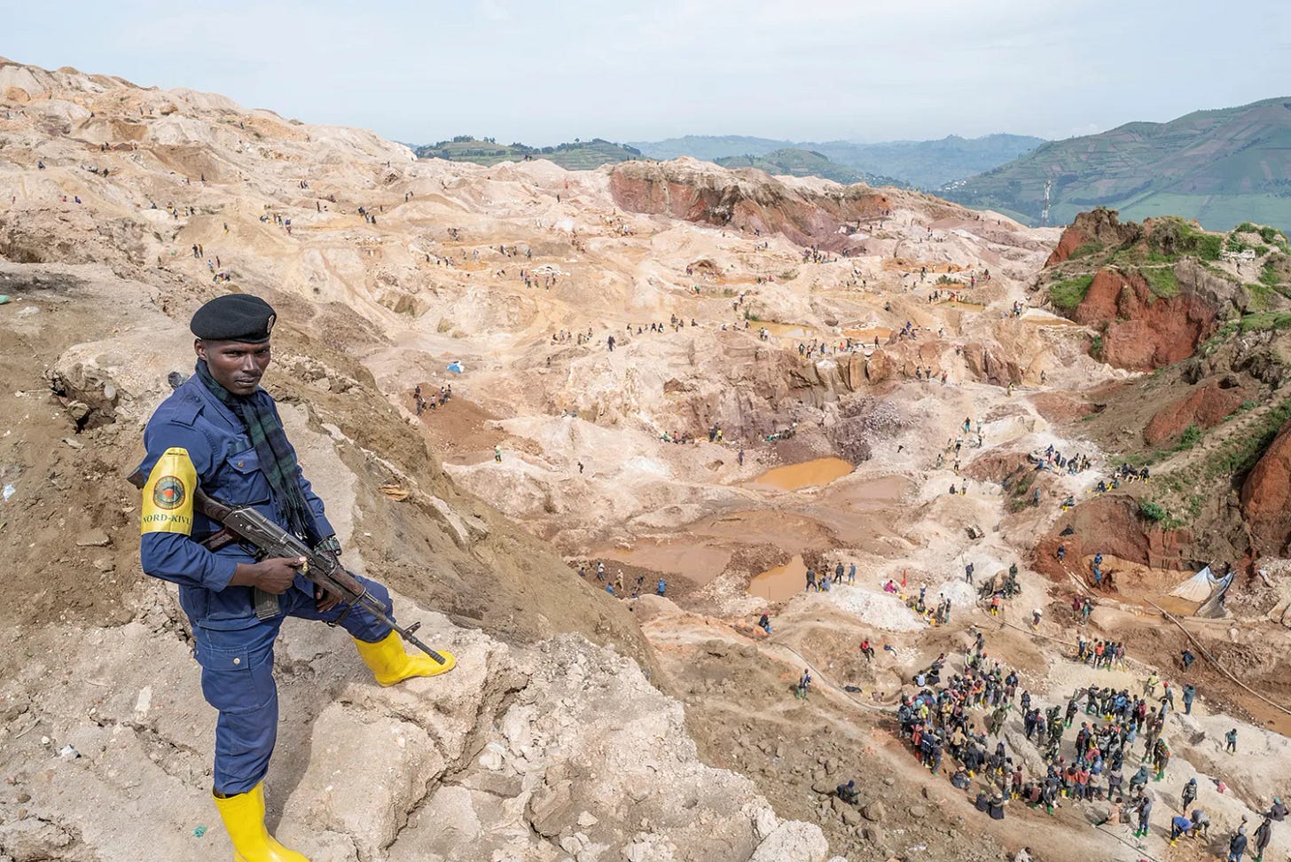 A police waring a uniform, beret and rubber boots holds a gun as he stands high on a hillside over a coltan mine. The barren landscape of a mine extends behind him and a cluster of workers are seen below.
