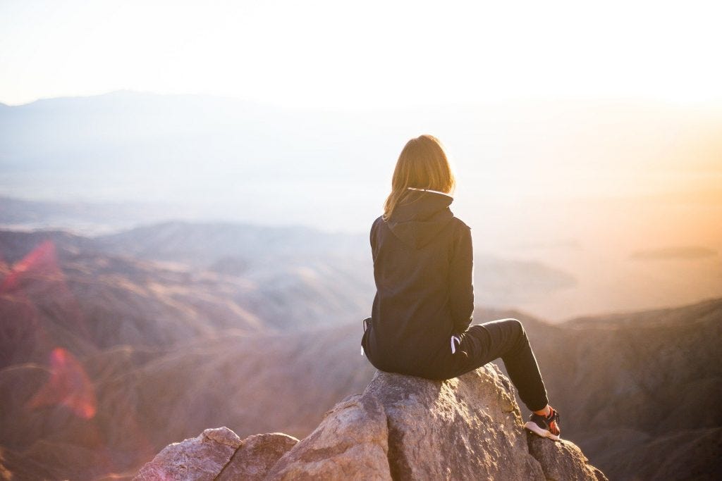 A woman sitting atop a rock at the top of a mountain overlooking other mountain vistas with a bright white obscuring part of the horizon. A woman sitting atop a rock at the top of a mountain overlooking other mountain vistas with a bright white obscuring part of the horizon.