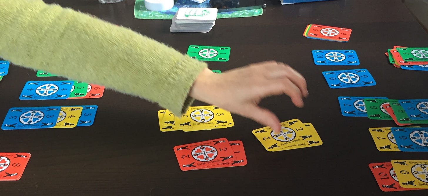 An arm stretches across a table to place a brightly coloured playing card on a stack. The table is strewn with many piles of cards.