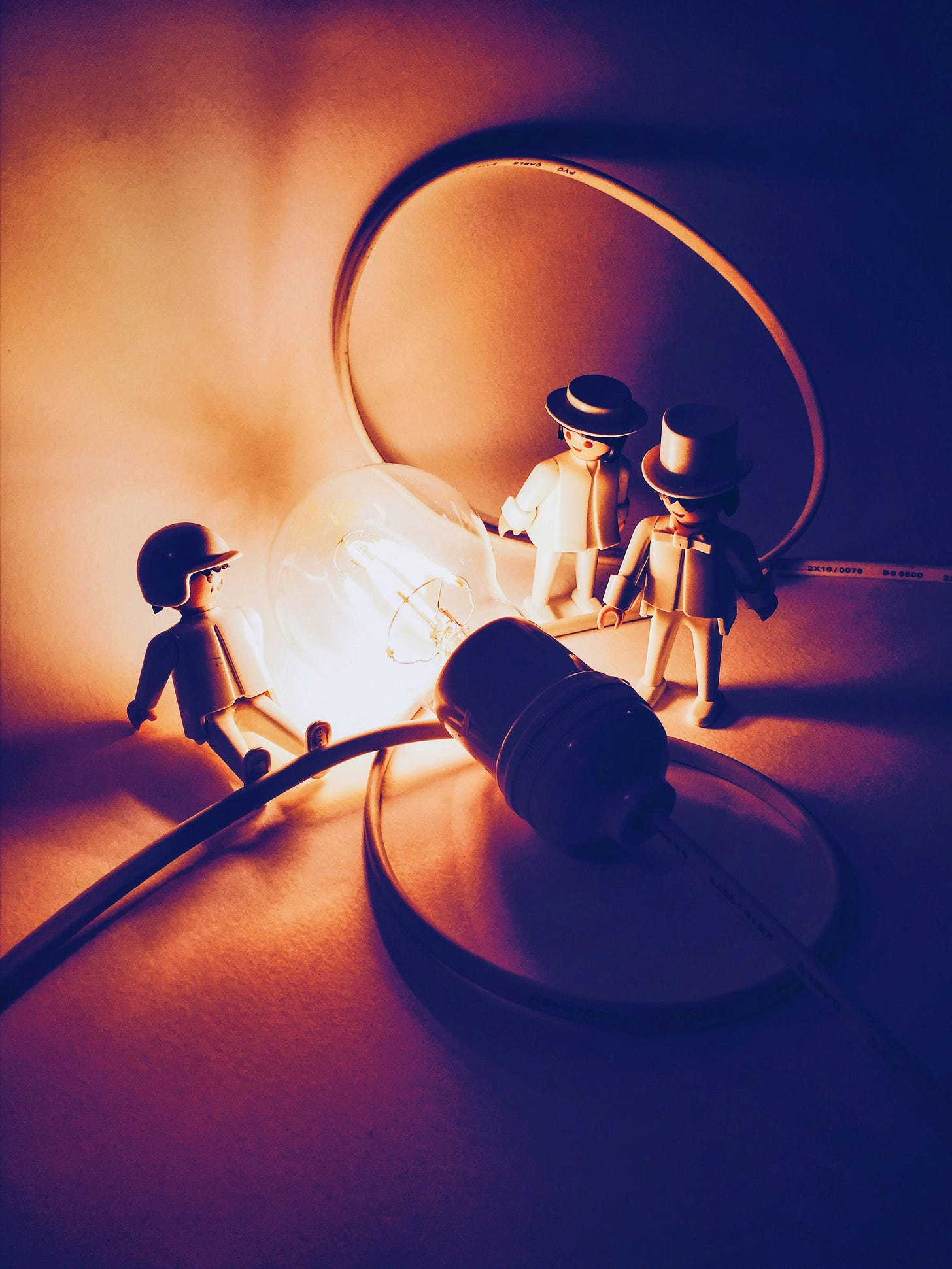 Three toy figures standing around a glowing light bulb on the floor, surrounded by loose electrical cables in a warm, dimly lit scene.