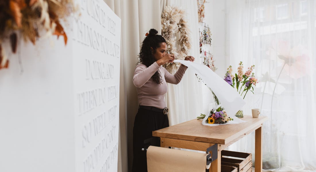 A woman standing over a wooden table, holding a sheet of paper over a bouquet, with a wreath and curtain behind her