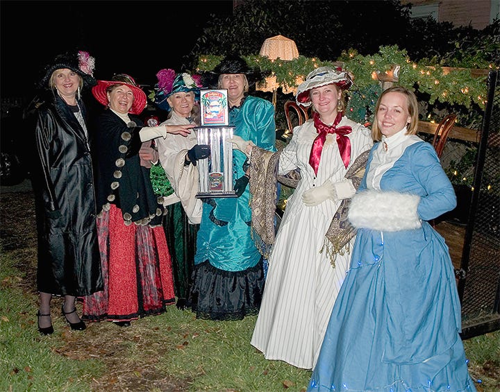 Nighttime photo of the six tour committee women dressed in vintage clothing, after the Christmas parade, and holding up the trophy they won for their float.