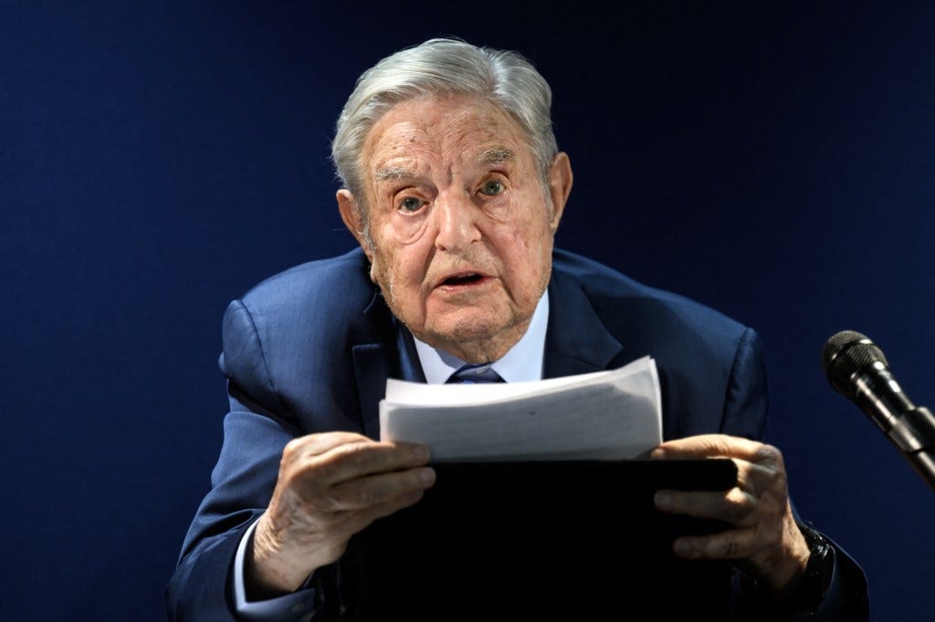 George Soros addressing an assembly, holding papers in his hands, with a microphone to his right. George Soros addressing an assembly, holding papers in his hands, with a microphone to his right.