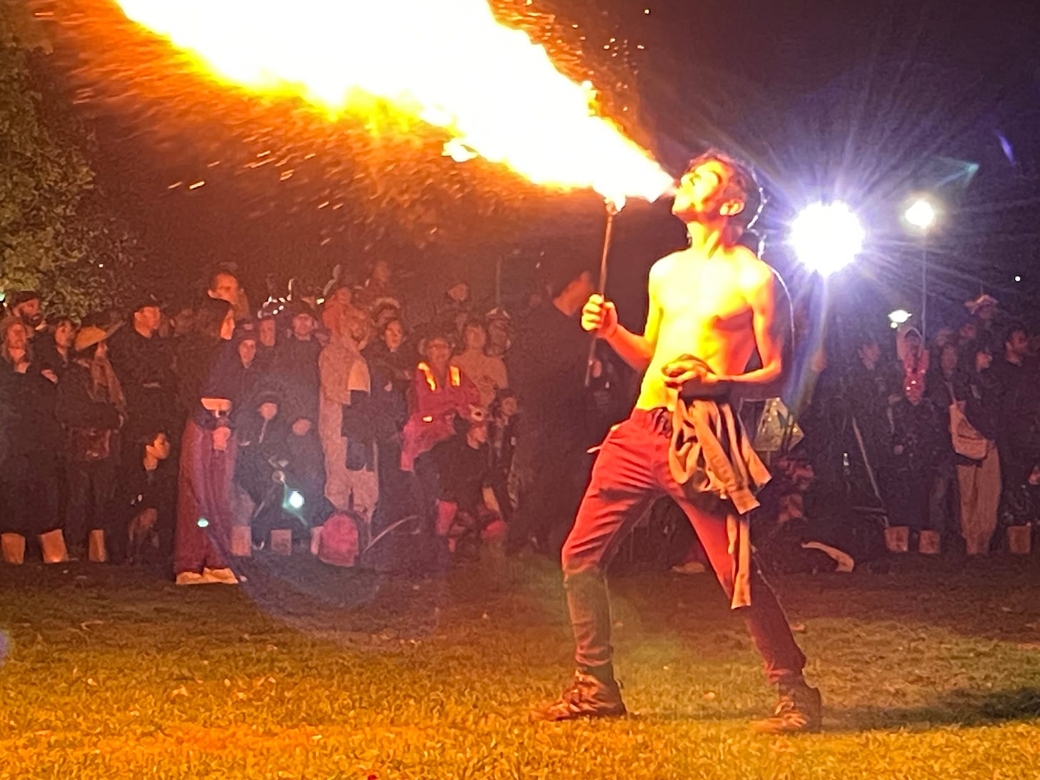 A shirtless man in an open space surrounded by a circle of onlookers as he breathes out a terrific, impressive gust of fire. A shirtless man in an open space surrounded by a circle of onlookers as he breathes out a terrific, impressive gust of fire.