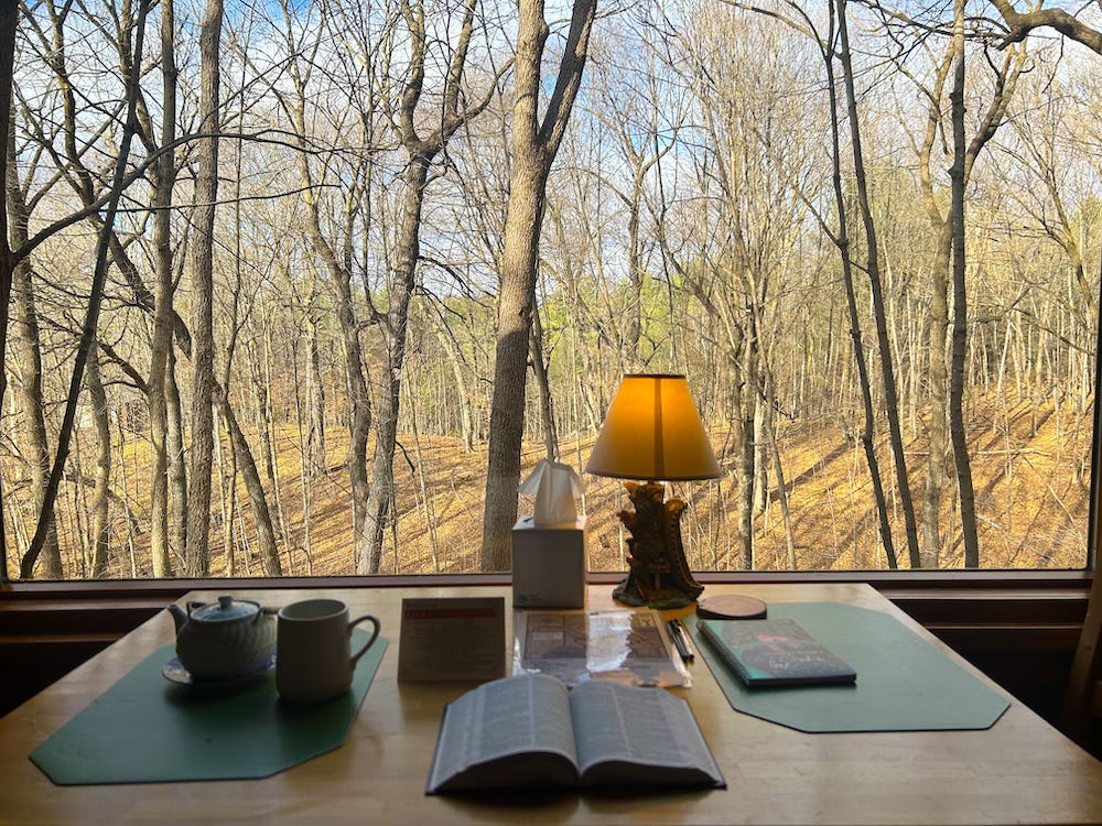 Picture shows a picture frame window with barren trees and a blue sky peeking out in the background. On the table is a Bible, lamp, tea pot, and mug. 