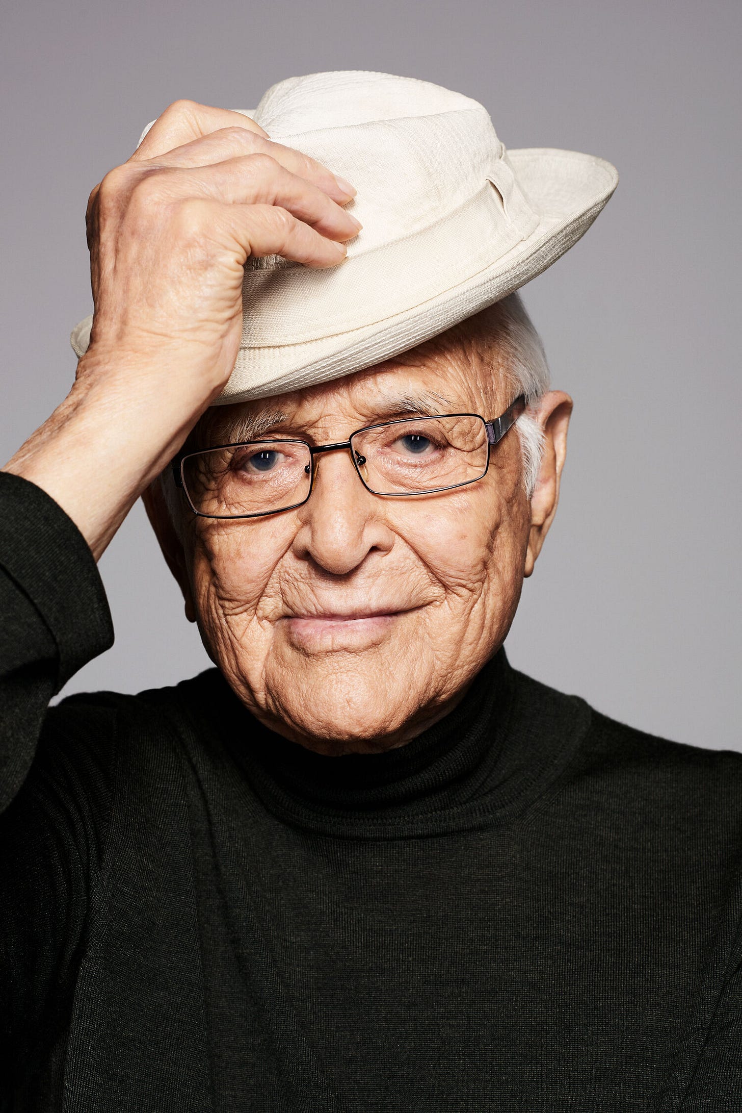Headshot of TV producer and writer Norman Lear. He is wearing glasses and a black turtleneck and looking into the camera. His right hand reaches up to his hat, which he tips slightly