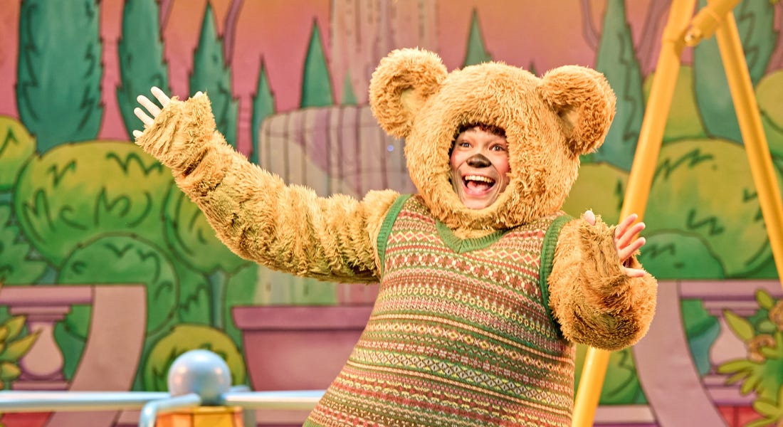 A man is jumping centre stage, his feet off the ground. He is wearing a fluffy teddy bear costume, his face is visible in a friendly smile but the rest of him is inside the costume. The bear has a green knitted sweater, and he is wearing orange trainers. Behind him the youth chorus are all dressed as childhood toys: ballerinas, clowns and nutcrackers. They’re also jumping in the air. The backdrop depicts the inside of a nursery.