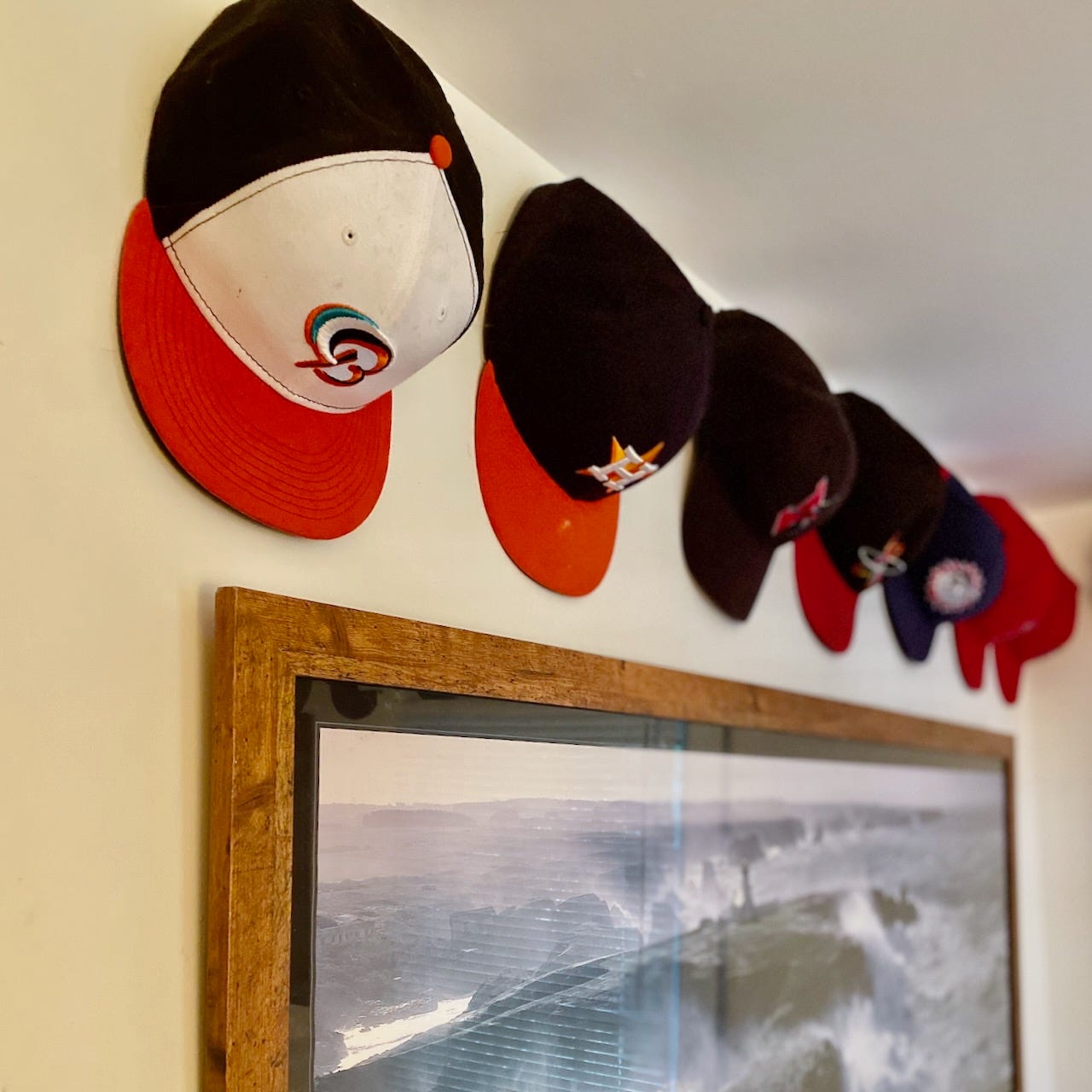 Rows of baseball caps mounted along the top of a bedroom wall, above a framed panoramic photograph.