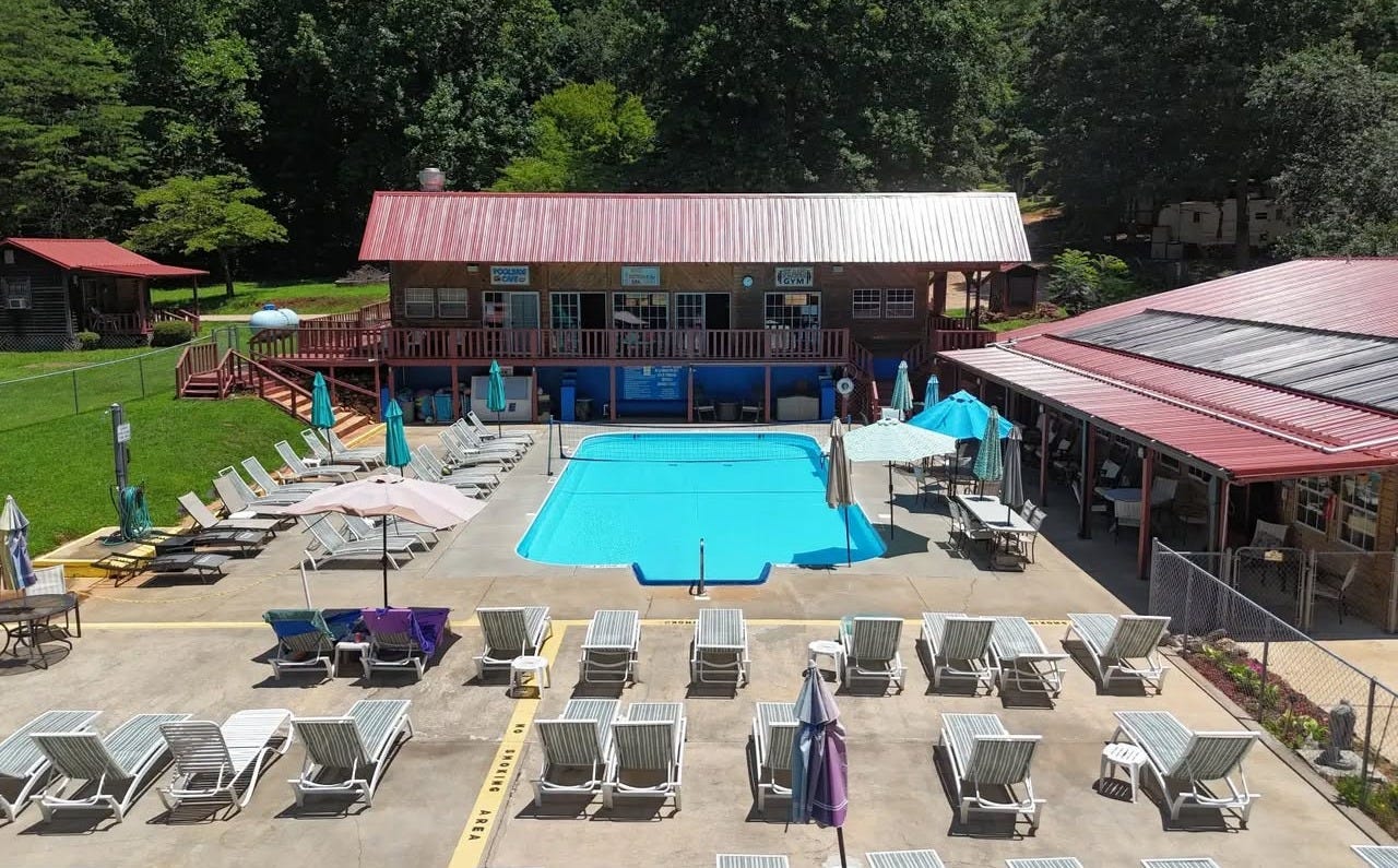 Aerial view of Serendipity Sun Club's swimming pool, surrounded by rows of lounge chairs and umbrellas, with a wood-and-red-roof clubhouse and a covered pavilion behind it, ringed by green north Georgia forest. Aerial view of Serendipity Sun Club's swimming pool, surrounded by rows of lounge chairs and umbrellas, with a wood-and-red-roof clubhouse and a covered pavilion behind it, ringed by green north Georgia forest.