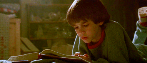 Gif dal film "La storia infinita" con un bambino sdraiato in una soffitta mentre legge un libro e poi lo chiude un attimo per guardare la copertina arancione su cui c'è uno stemma in rilievo di un uroboro, il serpente che si divora la coda.