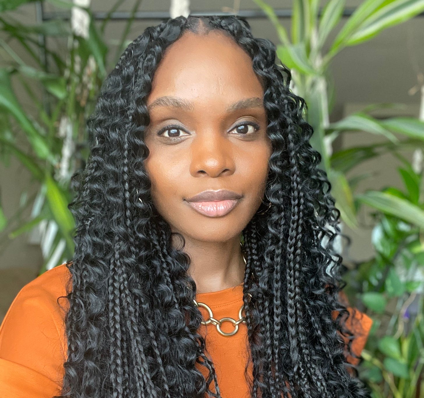 A head-and-shoulders portrait shows a person with long braided and curly hair wearing an orange top and a gold circular necklace. The subject faces the camera with a neutral expression, and the background contains large green indoor plants with soft natural lighting, producing a calm and professional atmosphere.