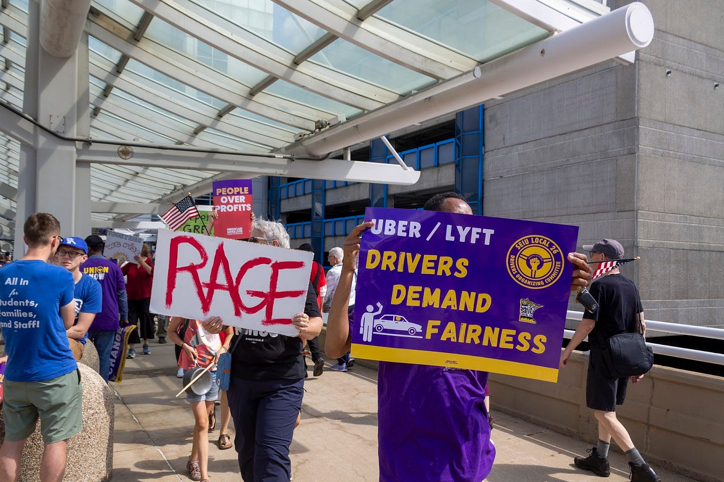 two people hold up signs, one with red text and the word “Rage” another with purple and yellow reading “Uber/Lyft Drivers Demand Fairness" two people hold up signs, one with red text and the word “Rage” another with purple and yellow reading “Uber/Lyft Drivers Demand Fairness"