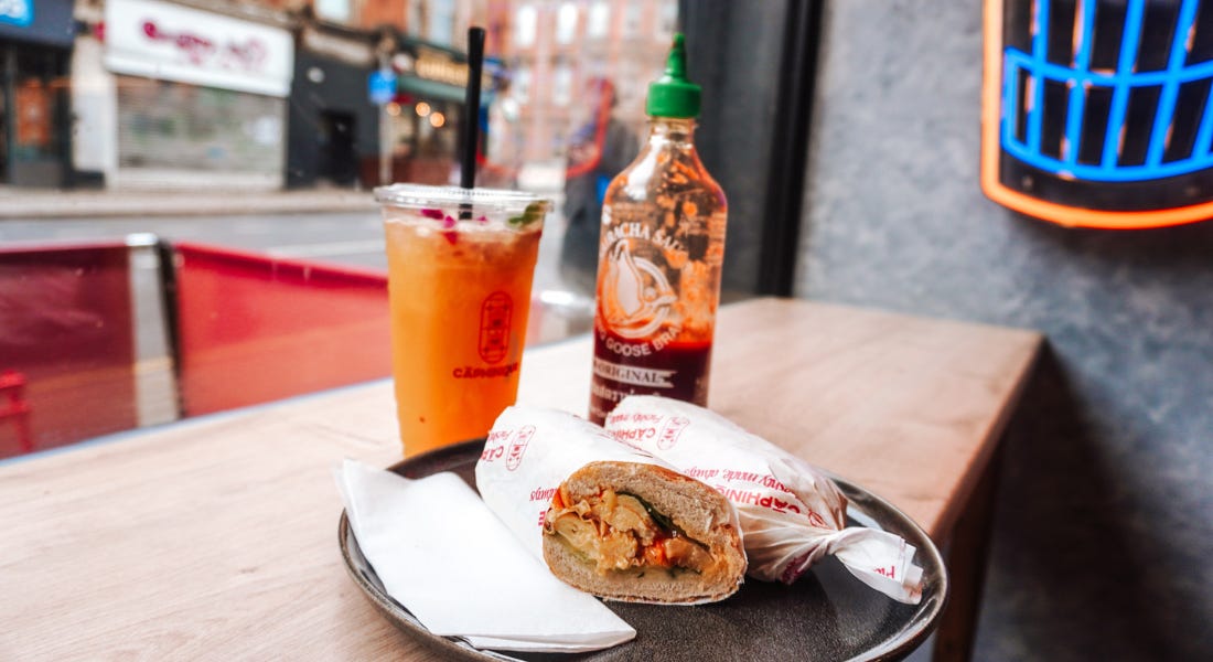A banh mi wrap with an orange drink and sriracha bottle next to it, and a street scene behind it